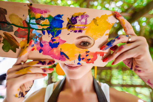 Smiling woman artist holding a paint-covered palette over her face, symbolizing creativity, emotional expression, and the healing power of art therapy